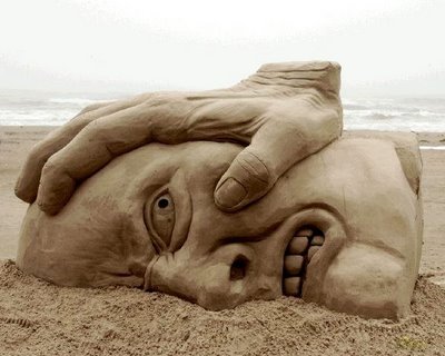 Gran escultura de arena en una playa. Representa un rostro furioso de hombre hundido en la arena por una mano. 